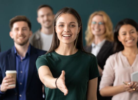 Friendly young company woman representative holds out her hand for handshake welcoming customer smiling looking at camera posing together with diverse colleagues, sales manager greeting client concept